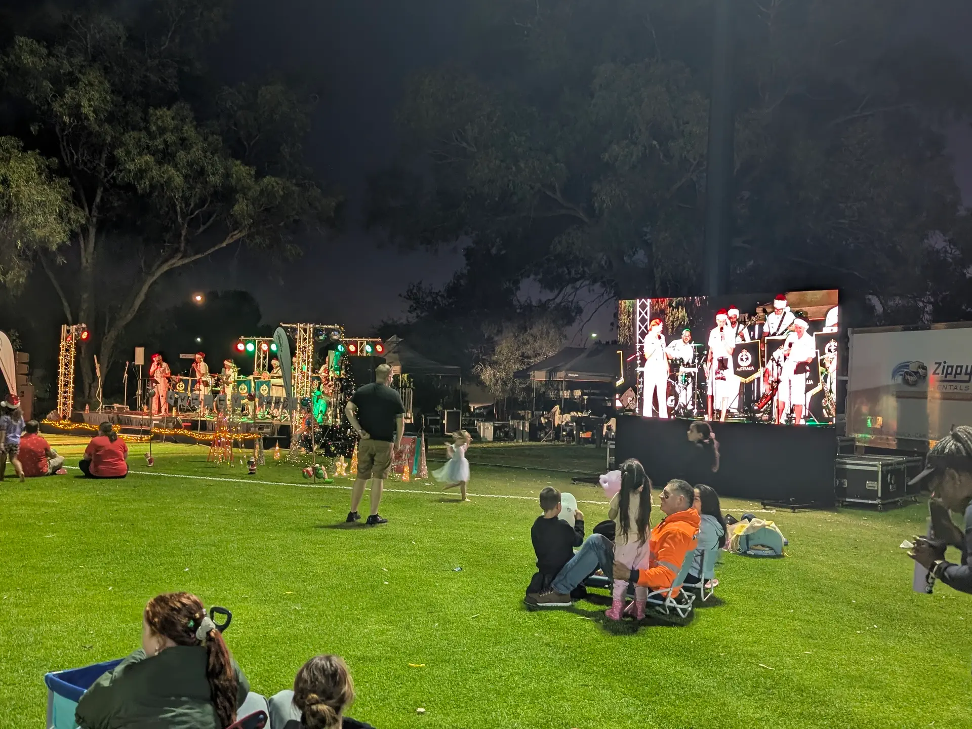 Families on a lawn at night watching an LED screen relay the carols stage