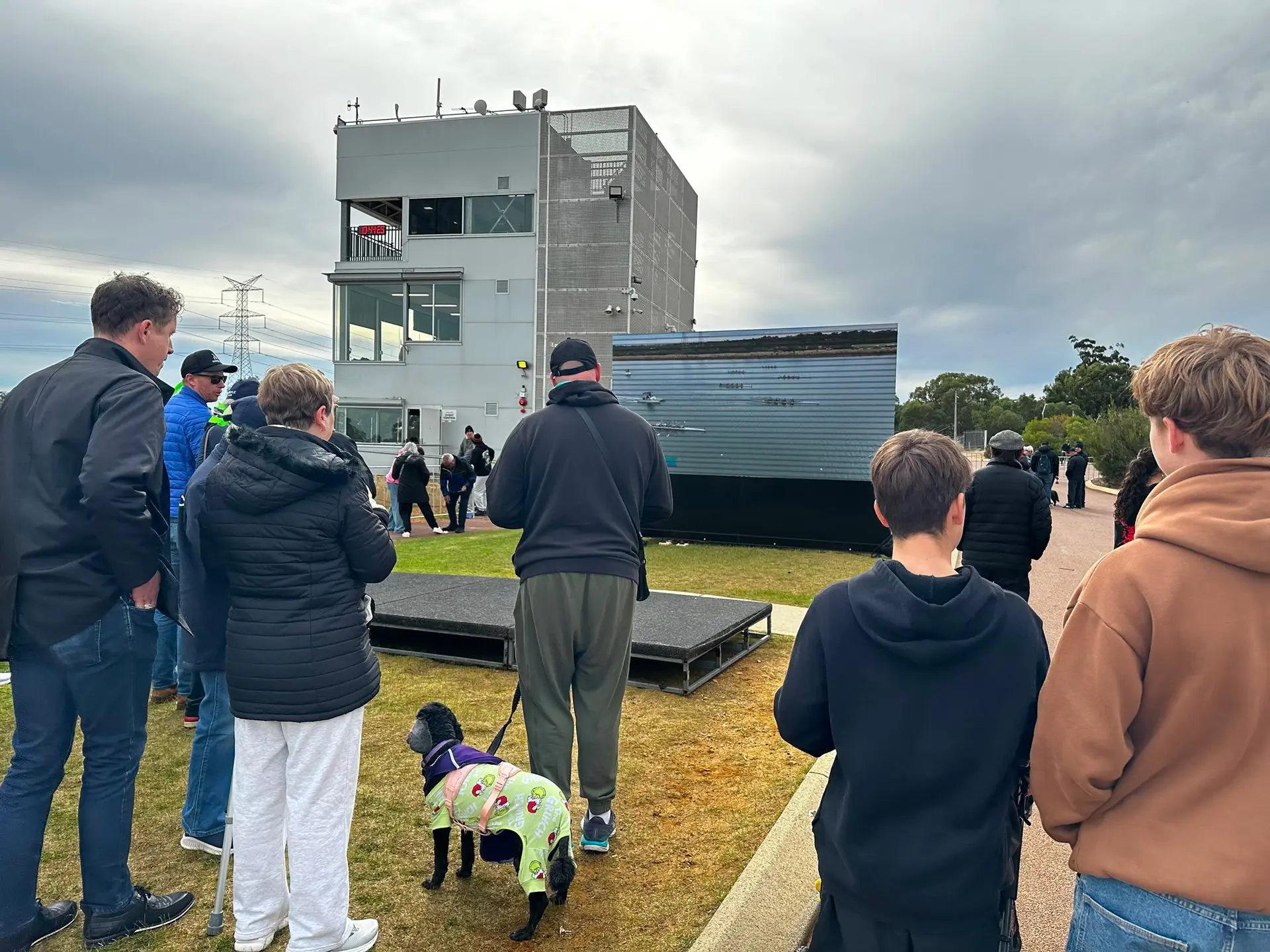 Spectators watching an LED screen beside the river at a school rowing regatta